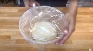 Person covering dough in a bowl with plastic wrap on a wooden countertop.